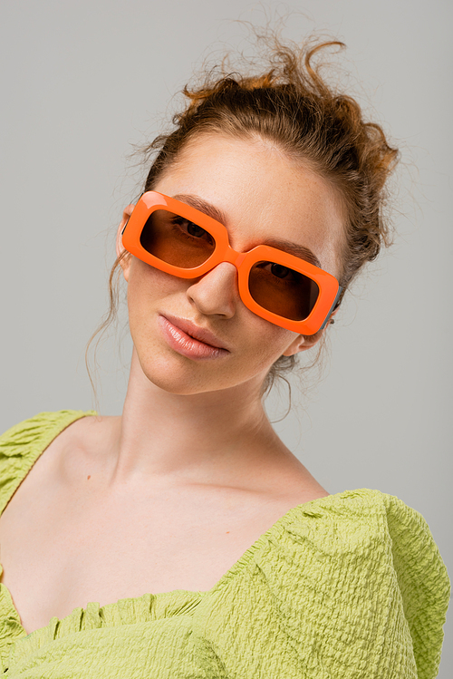 Portrait of young redhead woman in green blouse and modern sunglasses looking at camera while standing isolated on grey background, trendy sun protection concept, fashion model