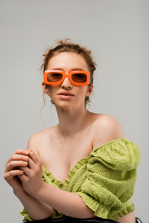 Portrait of stylish young red haired woman in sunglasses and green blouse with naked shoulders posing while standing isolated on grey background, trendy sun protection concept, fashion model