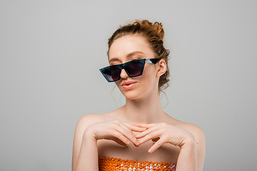 Young red haired woman in top with orange sequins and naked shoulders posing in sunglasses and standing isolated on grey background, trendy sun protection concept, fashion model