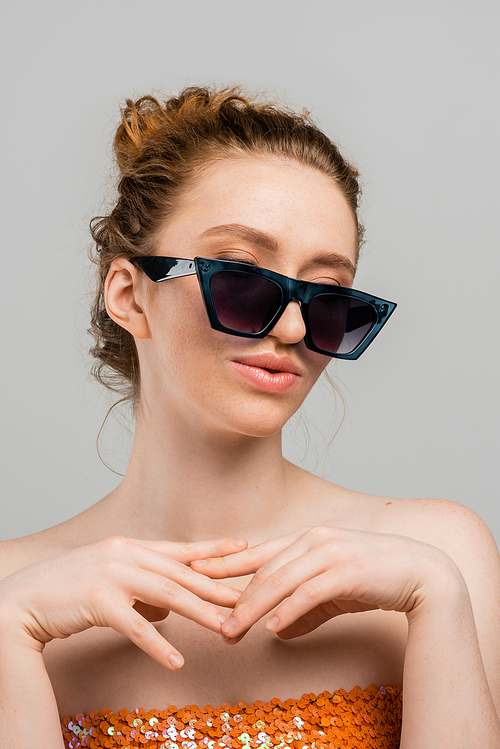 Portrait of young redhead woman with freckles wearing top with sequins and stylish sunglasses while standing isolated on grey background, trendy sun protection concept, fashion model