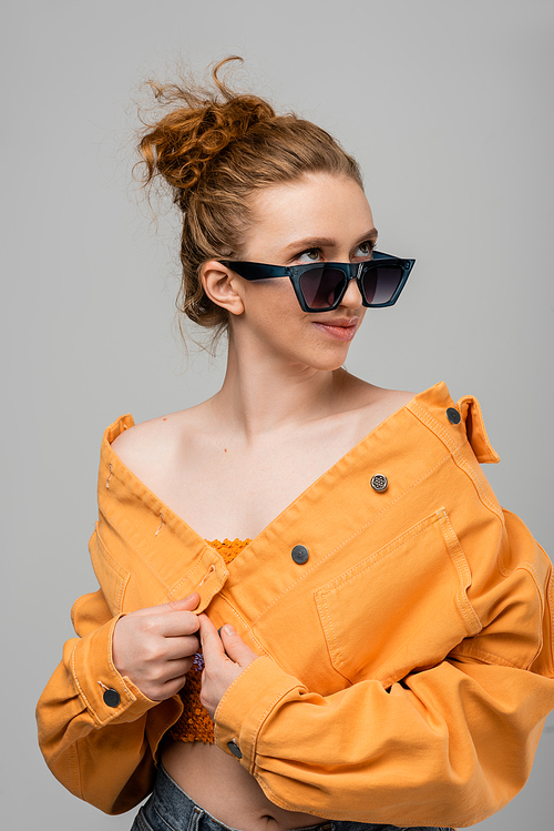 Smiling and trendy young redhead woman in sunglasses and orange denim jacket looking away while posing isolated on grey background, trendy sun protection concept, fashion model
