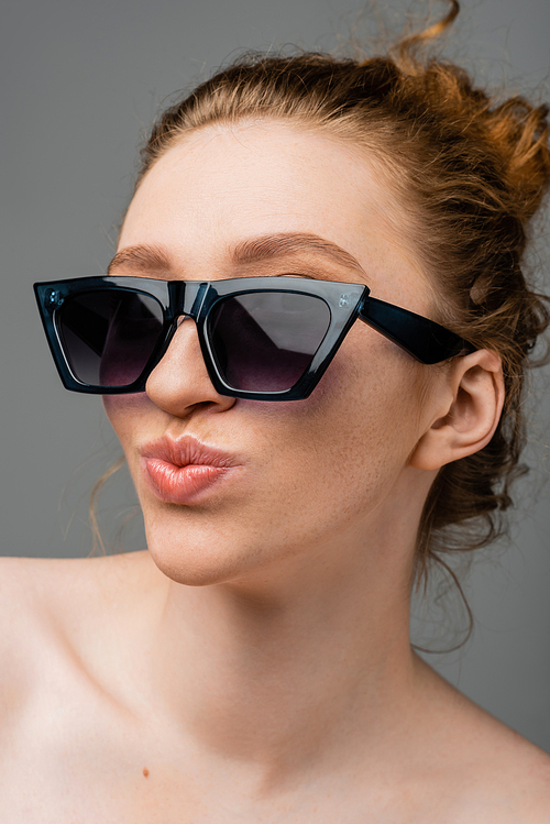 Close up view of young redhead and freckled woman in stylish sunglasses with naked shoulders pouting lips and standing isolated on grey background, trendy sun protection concept, fashion model