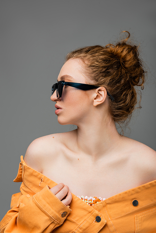 Side view of young red haired woman in sunglasses and orange denim jacket with naked shoulders looking away isolated on grey background, trendy sun protection concept, fashion model