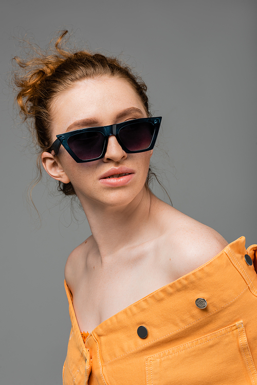 Portrait of red haired and freckled model in sunglasses and orange denim jacket posing and standing isolated on grey background, trendy sun protection concept, fashion model