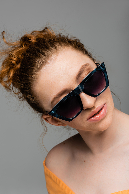 Portrait of fashionable young and red haired woman with natural makeup wearing sunglasses while standing isolated on grey background, trendy sun protection concept, fashion model