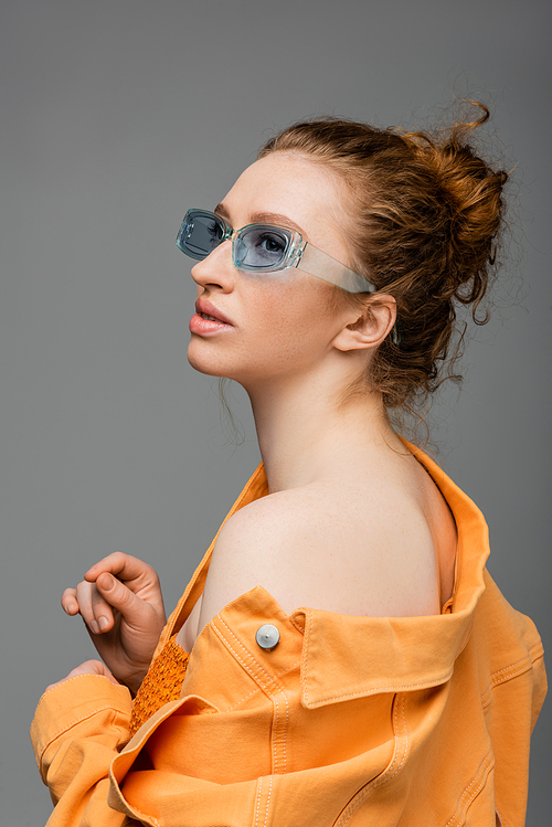 Fashionable young red haired woman in sunglasses and orange denim jacket with naked shoulder looking away and standing isolated on grey background, trendy sun protection concept, fashion model
