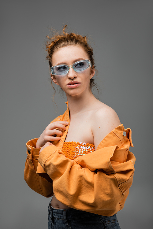 Stylish young red haired woman in sunglasses, top with sequins and orange denim jacket with naked shoulder standing isolated on grey background, trendy sun protection concept, fashion model