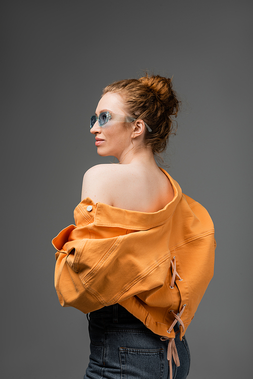 Stylish young red haired woman in sunglasses and orange denim jacket with naked shoulder looking away while standing isolated on grey background, trendy sun protection concept, fashion model
