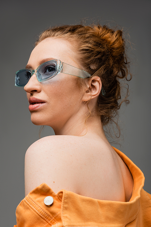 Portrait of red haired and freckled young woman in sunglasses and orange denim jacket with naked shoulder standing isolated on grey background, trendy sun protection concept, fashion model