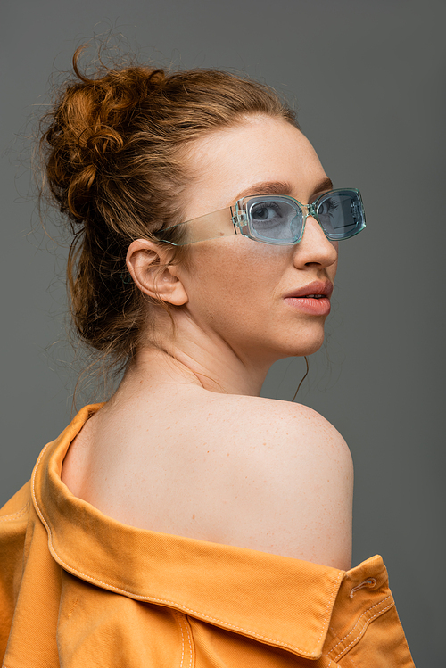 Modern red haired woman in blue sunglasses and orange denim jacket with naked shoulder looking away and standing isolated on grey background, trendy sun protection concept