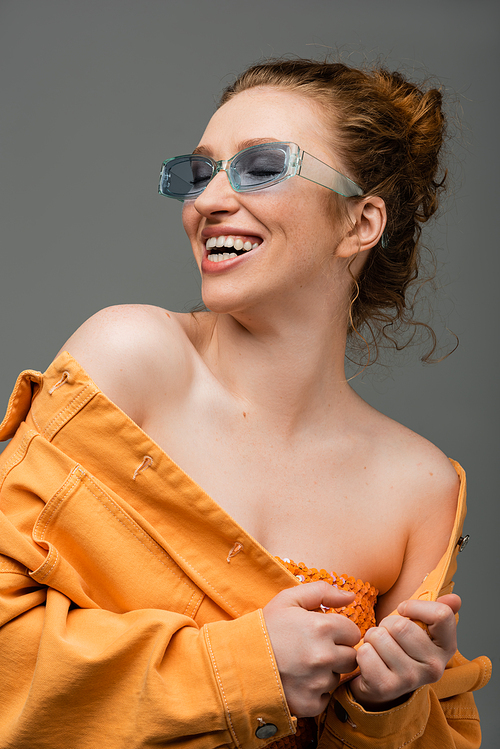 Cheerful and stylish red haired woman in blue sunglasses and orange denim jacket with naked shoulders laughing isolated on grey background, trendy sun protection concept