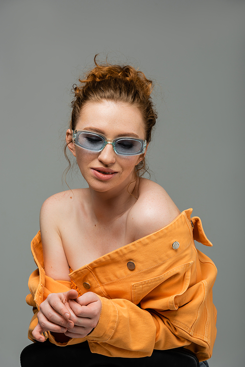Smiling and stylish red haired woman in sunglasses posing in orange denim jacket with naked shoulders isolated on grey background, trendy sun protection concept, fashion model