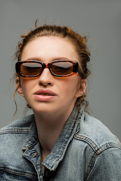 Portrait of stylish young redhead woman with freckles posing in sunglasses and denim jacket and standing isolated on grey background, trendy sun protection concept, fashion model