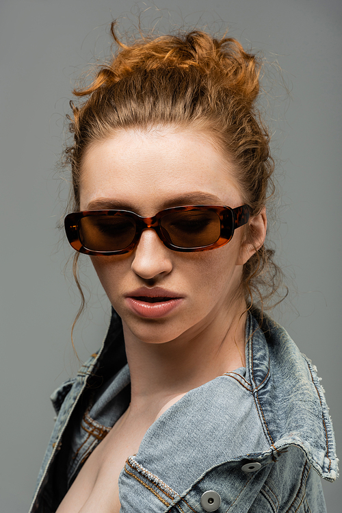 Stylish and confident young redhead model with natural makeup in sunglasses posing in denim jacket and standing isolated on grey background, trendy sun protection concept, fashion model