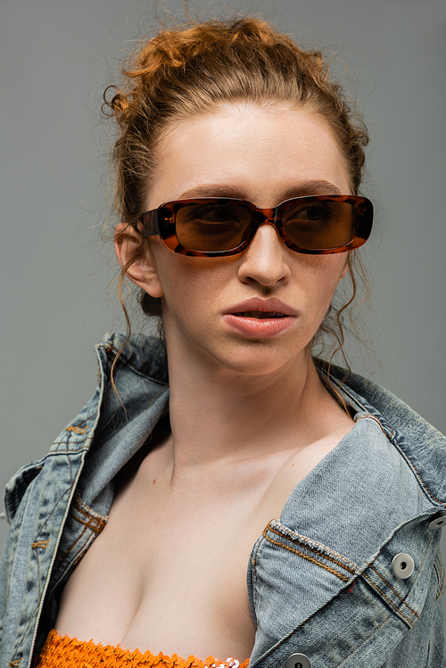Confident young red haired woman with natural makeup wearing sunglasses and denim jacket and looking away isolated on grey background, trendy sun protection concept, fashion model