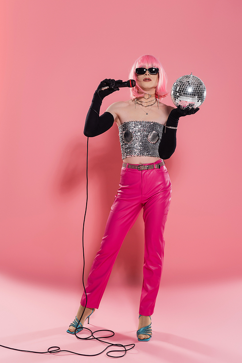 Full length of trendy drag queen in sunglasses and gloves holding microphone and disco ball on pink background