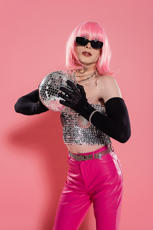 Fashionable drag queen in sunglasses and gloves holding disco ball on pink background