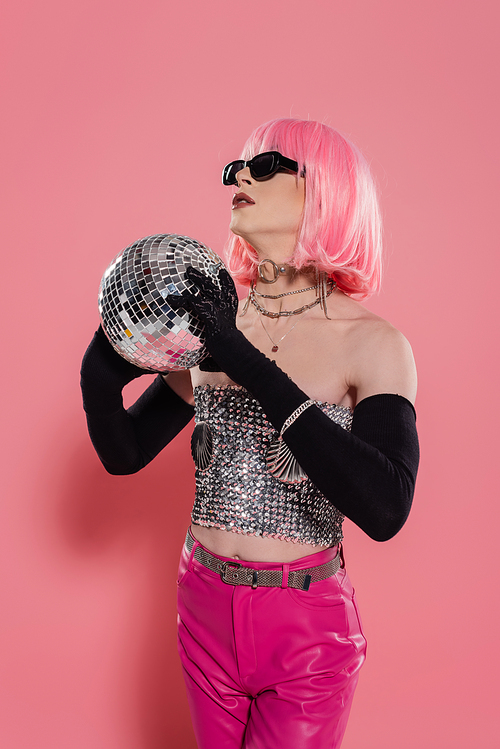 Stylish transgender person in sunglasses and gloves holding disco ball on pink background