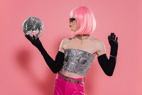 Side view of trendy drag queen in sparkling top and gloves holding disco ball on pink background