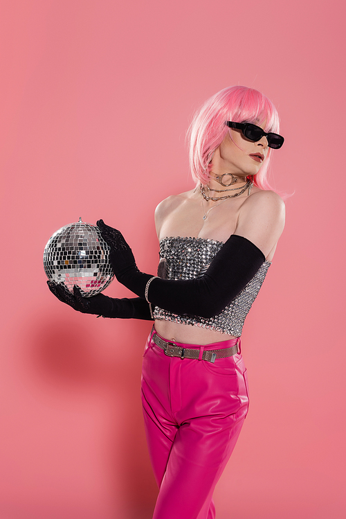 Fashionable drag queen in sunglasses and silver top holding disco ball on pink background