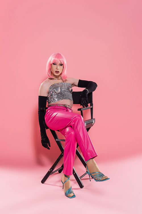 Fashionable drag queen in shiny top sitting on chair on pink background