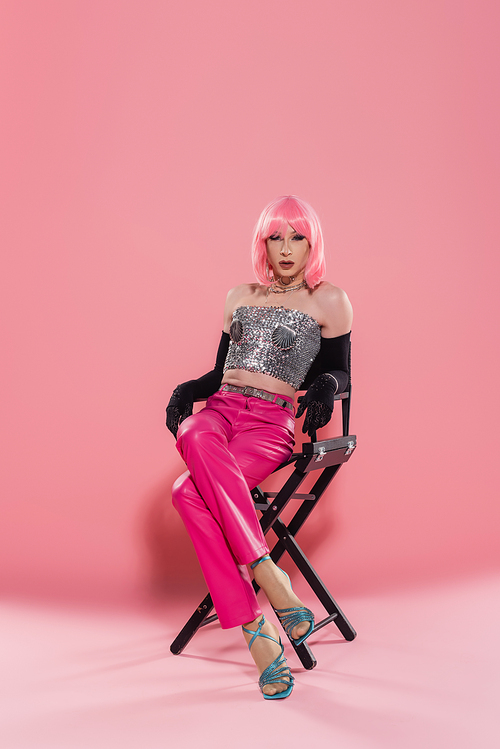Fashionable drag queen in top and gloves posing while sitting on chair on pink background