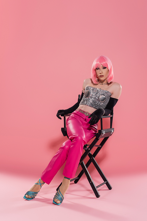 Stylish drag queen in top and wig sitting on chair on pink background
