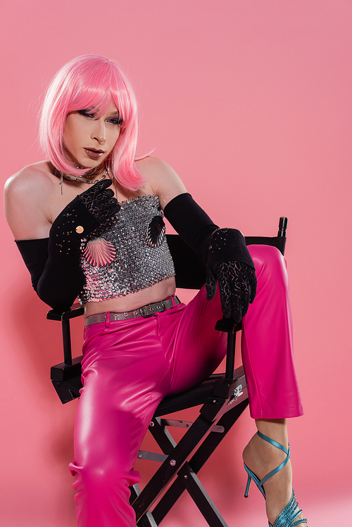 Fashionable drag queen in gloves and top sitting on chair on pink background