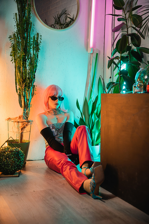 Stylish transgender person in sunglasses sitting on floor near plants at home