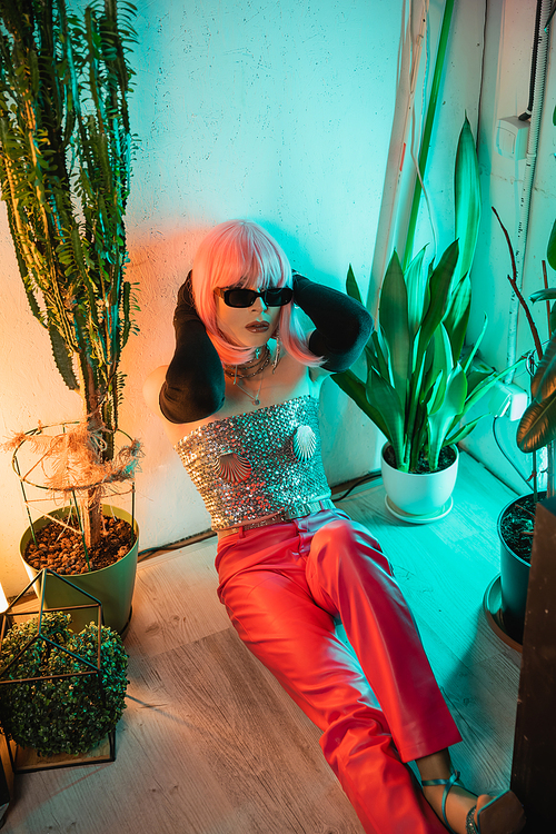 Trendy drag queen in pink wig and sunglasses sitting on floor near plants at home