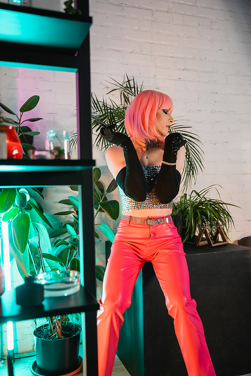 Side view of stylish drag queen in pink wig sitting on table with plant at home