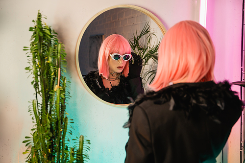 Trendy drag queen in wig and sunglasses standing near mirror and plants at home