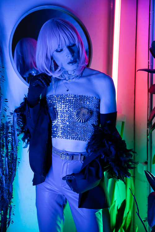 Trendy transgender person in wig and jacket standing near neon light at home