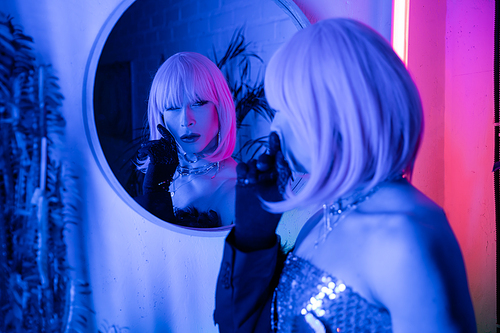 Fashionable drag queen in wig looking at mirror in neon light at home