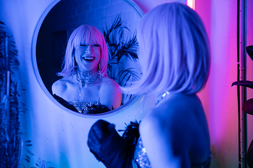 Cheerful drag queen in wig and top looking at mirror in neon light at home