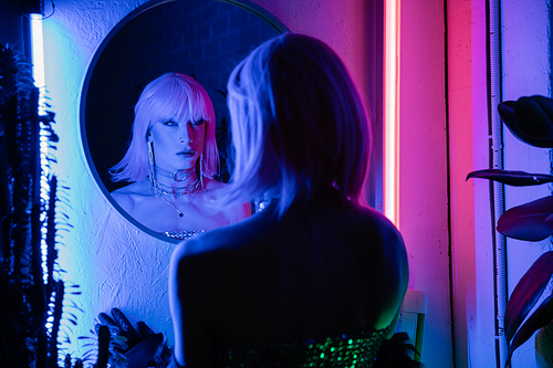 Blurred transgender person in wig and top looking at mirror near plants and neon light at home