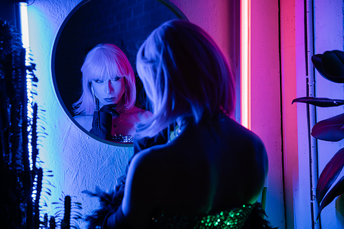Fashionable drag queen looking at mirror while standing near plants and neon light at home