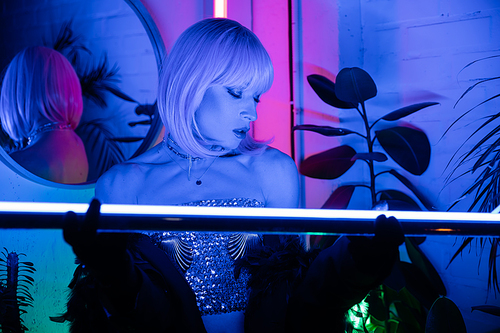 Trendy drag queen in wig and top holding neon lamp at home