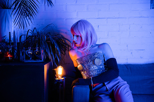 Drag queen in wig and top looking at lamp near plants at home