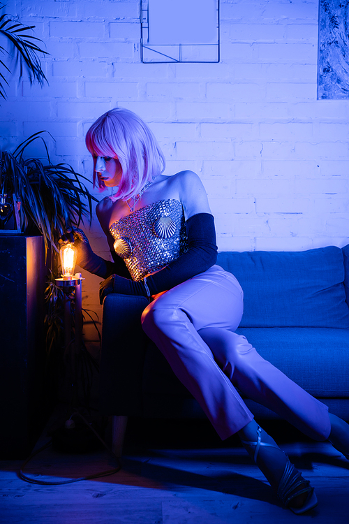 Stylish drag queen in gloves and top touching lamp while sitting on couch at home