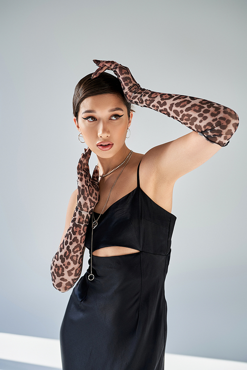 appealing asian woman with brunette hair and bold makeup posing in black strap dress and animal print gloves on grey background, spring style, modern fashion photography