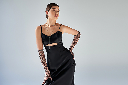 spring style, expressive asian woman with brunette hair and bold makeup standing with hand on hip in black elegant strap dress, animal print gloves and silver necklaces on grey background