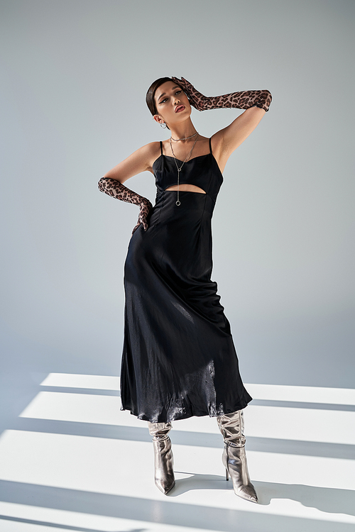 full length of glamour asian woman posing with hand on hip in black strap dress, animal print gloves and silver boots on grey background with lighting and shadows, spring style, fashion shoot