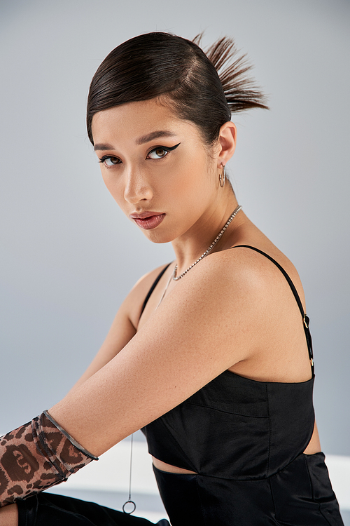 spring style asian woman with bold makeup, brunette hair and expressive gaze looking at . on grey background, black strap dress, silver necklaces, animal print glove, fashion shoot