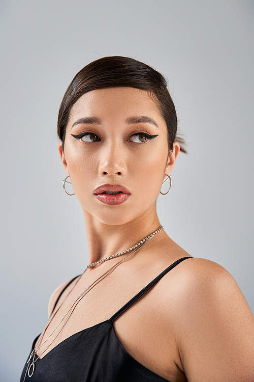 portrait of alluring asian woman in elegant black dress, silver necklaces and earrings, with bold makeup and brunette hair looking away on grey background, fashionable spring