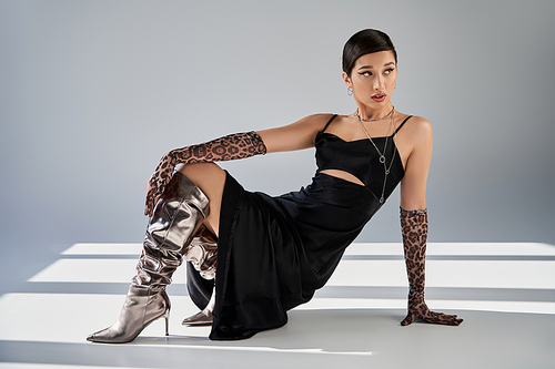 full length of trendy asian woman with bold makeup, in silver boots, black strap dress and animal print gloves posing and looking away on grey background with lighting and shadows, spring fashion