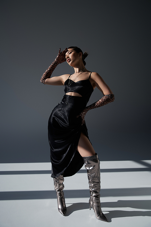 full length of alluring asian woman in silver boots, black fashionable dress and animal print gloves standing with hand on hip on dark grey background with lighting, trendy fashion concept