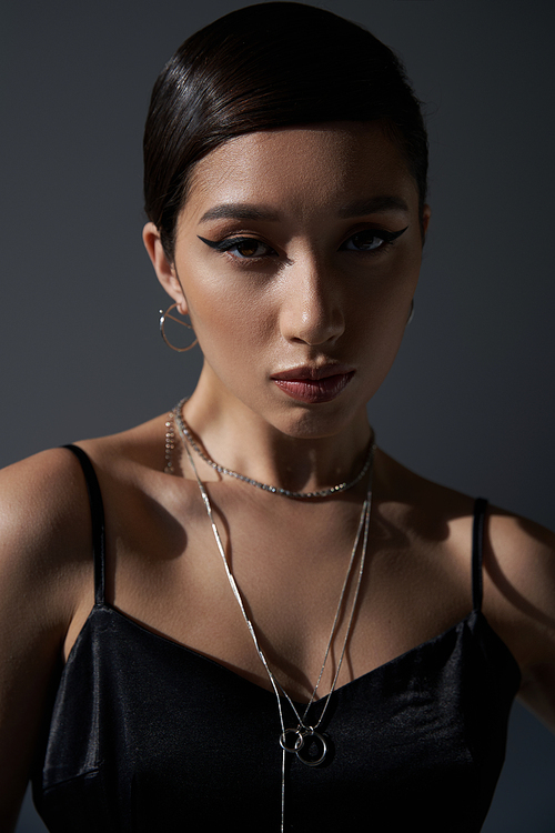 portrait of charming asian woman with bold makeup and brunette hair, in black strap dress and silver necklaces looking at . on dark grey background, generation z, spring style concept