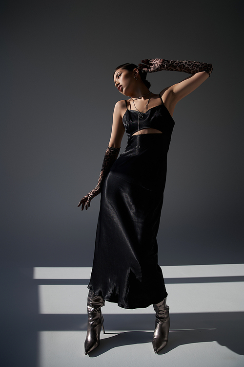 full length of graceful asian woman in black strap dress, animal print gloves and silver boots standing in stylish pose on dark grey background with lighting, spring fashion photography