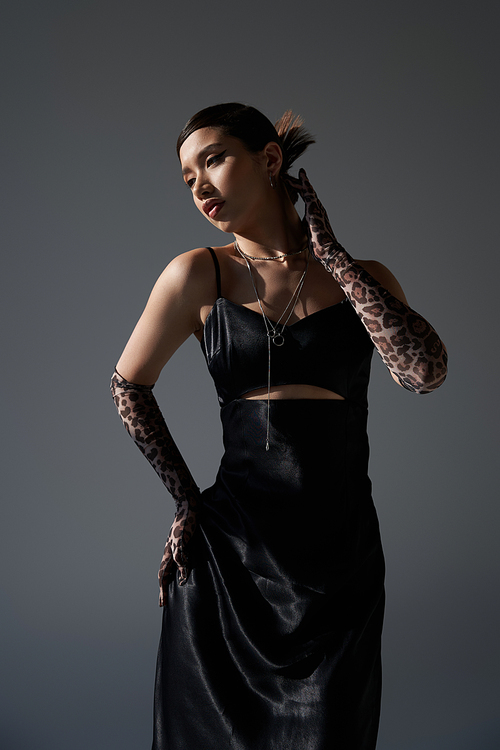 youthful fashion, stylish spring, sensual asian woman with brunette hair, in black elegant dress and animal print gloves posing with hand on hip on dark grey background